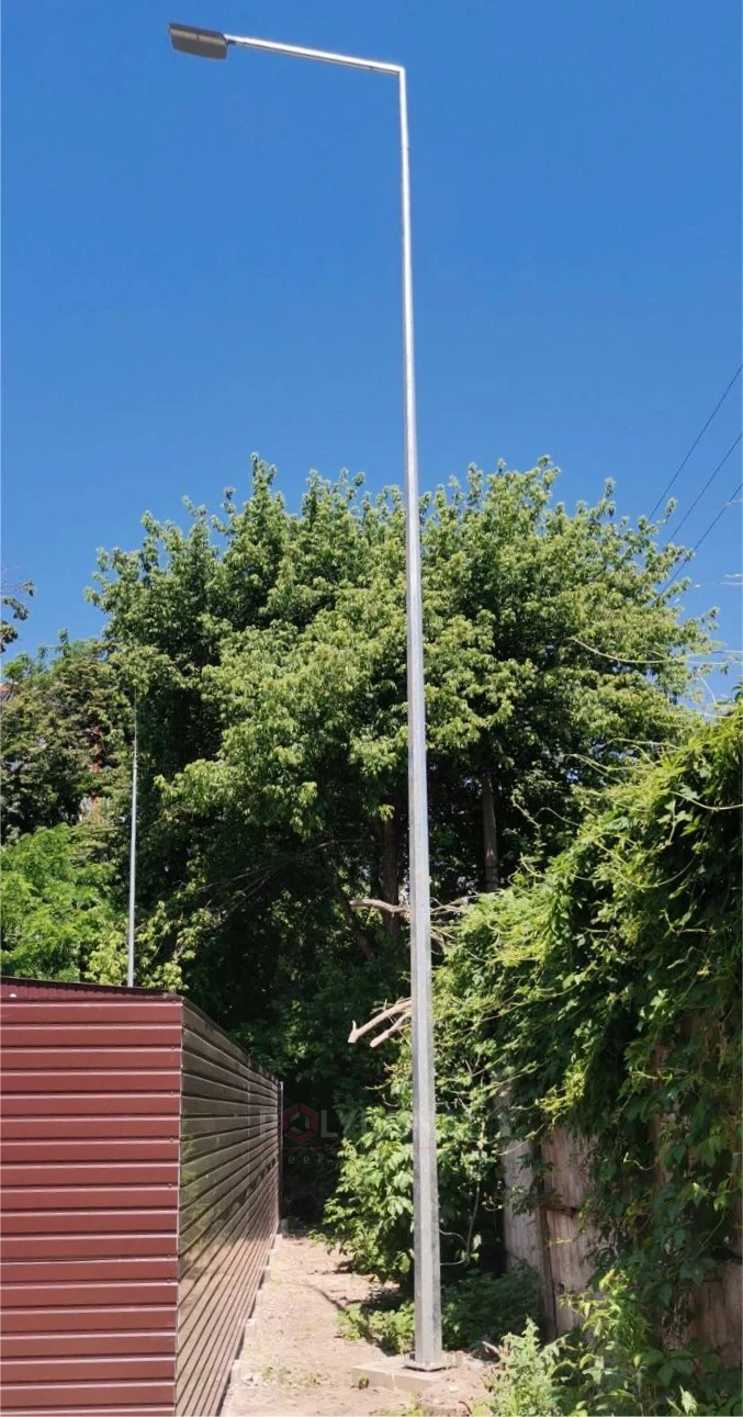PMO pole with a bracket-mounted luminaire between a fence and greenery